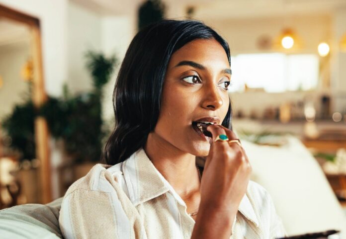 Home,,Woman,And,Eating,Chocolate,For,Relax,,Comfort,Food,And Home, woman and eating chocolate for relax, comfort food and hungry for sugar snack in living room. Delicious, sweets and person with dark cacao treat for confectionery candy, think and enjoy dessert